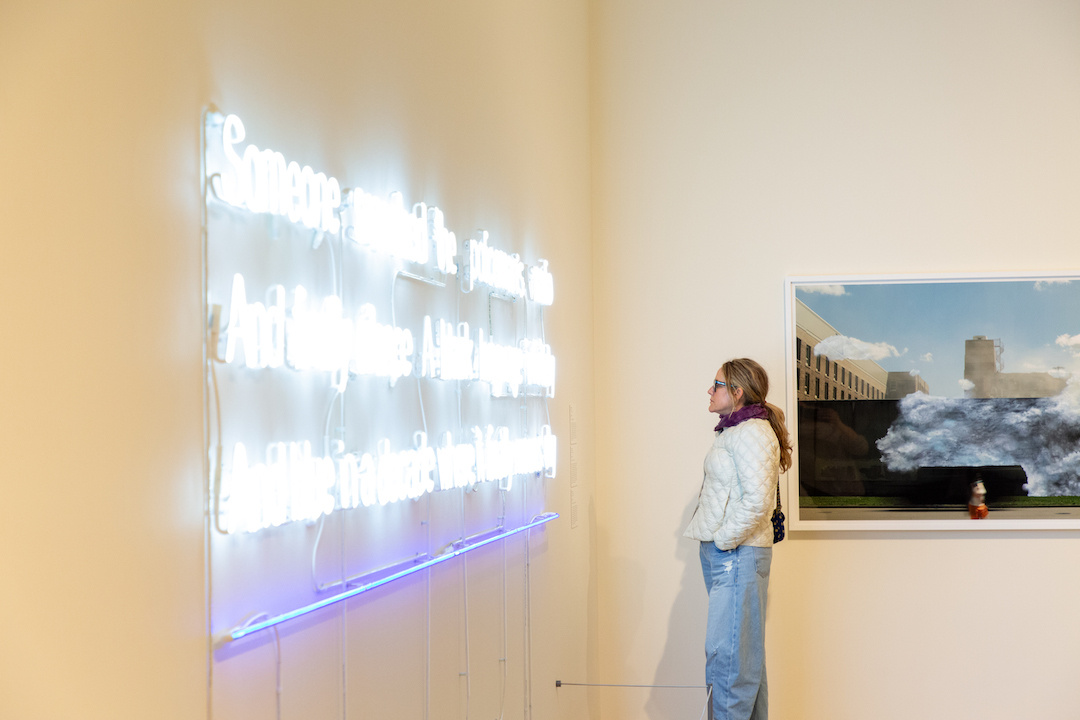 A woman in a white jacket and jeans observes a neon text art installation in a gallery