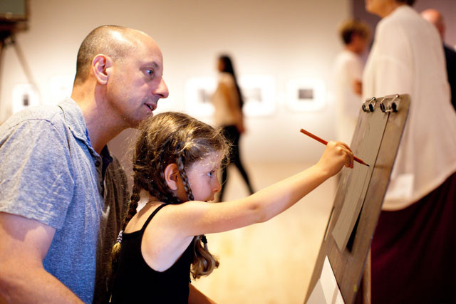 Sitting on her father’s lap, a young girl draws on an art easel in a gallery. People are in the background.