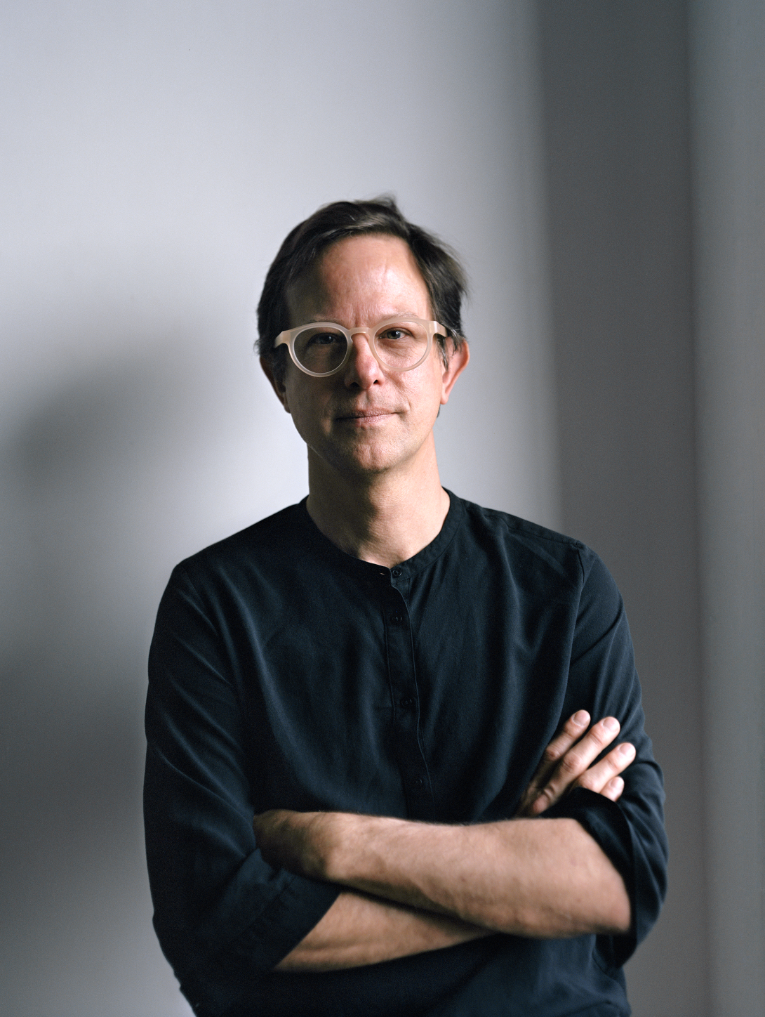 A photograph of a white man with arms folded and wearing white glasses. He looks directly at the camera and stands against a grey background. 