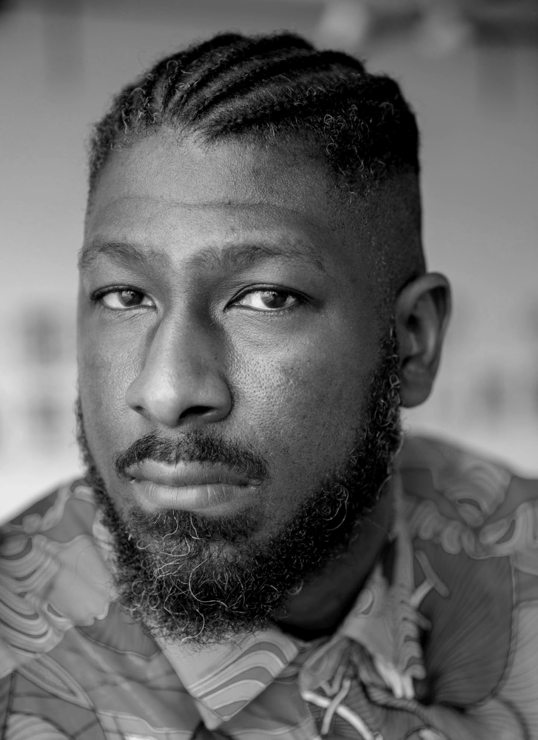 A black and white close up portrait of a Black man who looks directly at the camera. His lips are closed and his hair is braided. 