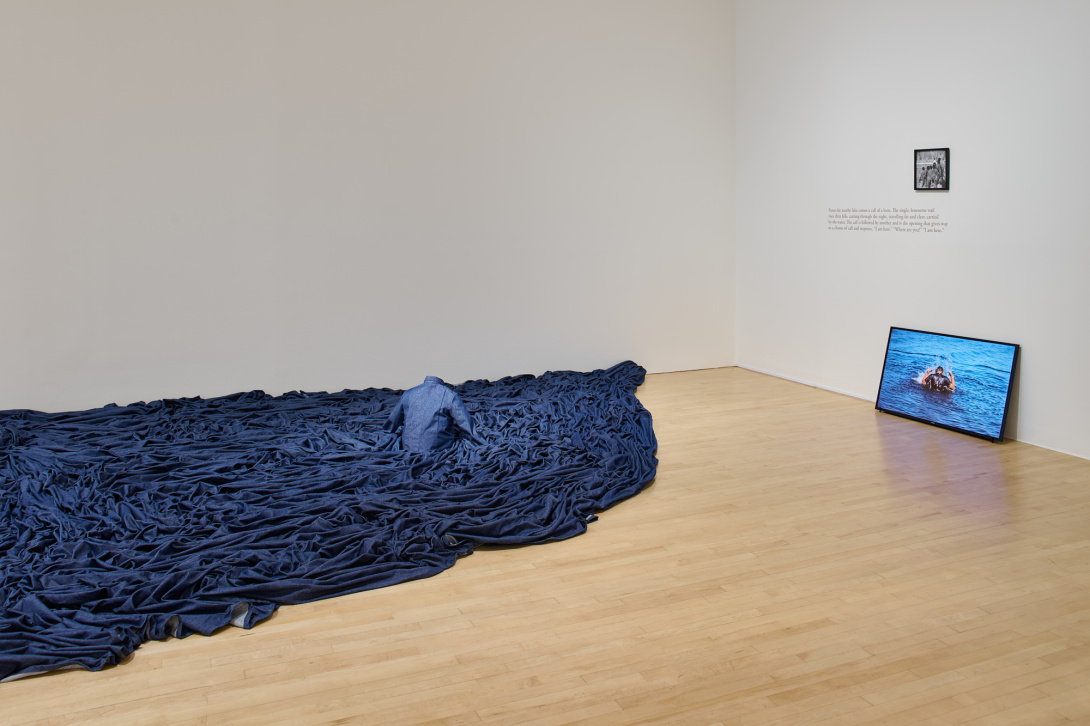 an installation of rippled denim fabric reminiscent of water with a headless figure emerging from it facing a flat screen monitor displaying a person in water with a vintage photograph above
