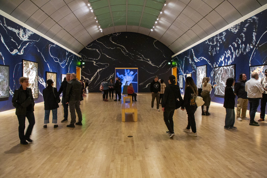 Visitors explore a dimly lit art gallery with a deep blue, celestial-themed mural, framed artworks on the walls, and a central video projection