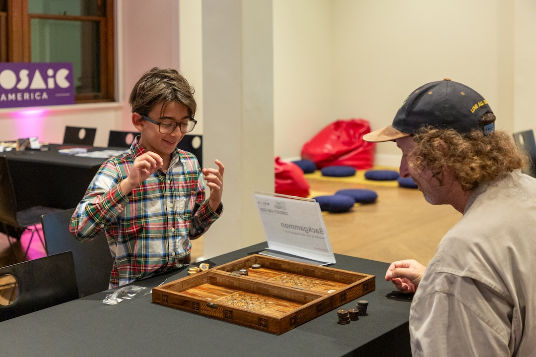 young boy with glasses cheerfully playing backgammon with a man with curly hair wearing a baseball cap