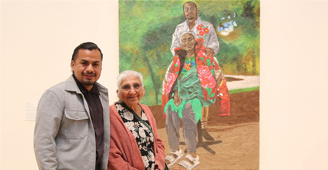 A smiling young man stands beside an older woman with gray braids, both in front of a large painted portrait of themselves.