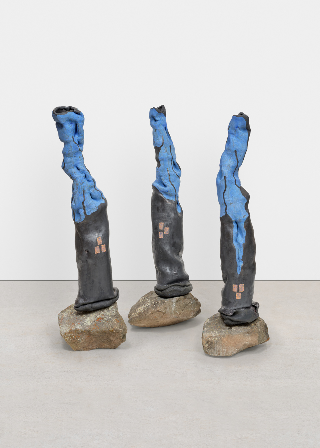 A sculpture comprised of three contorted pieces that each rest on large rocks. They each have a base of dark grey with sky blue paint dripping from the top down.