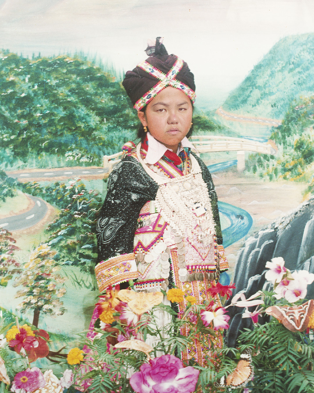 A young boy dressed in ceremonial clothing, surrounded by photos of colorful flowers with mountains and a river in the background.