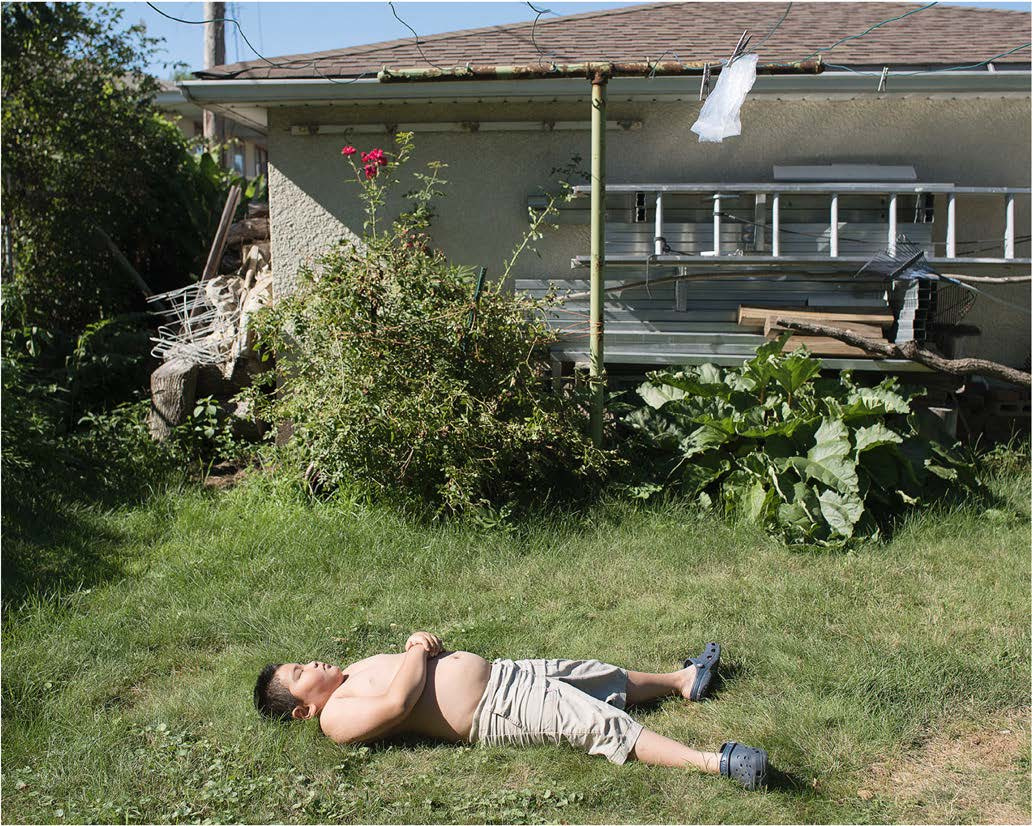 A young boy, shirtless in long shorts and shoes, lays on the grass. Behind him is a fence and a house. Flying above him is a plastic bag.