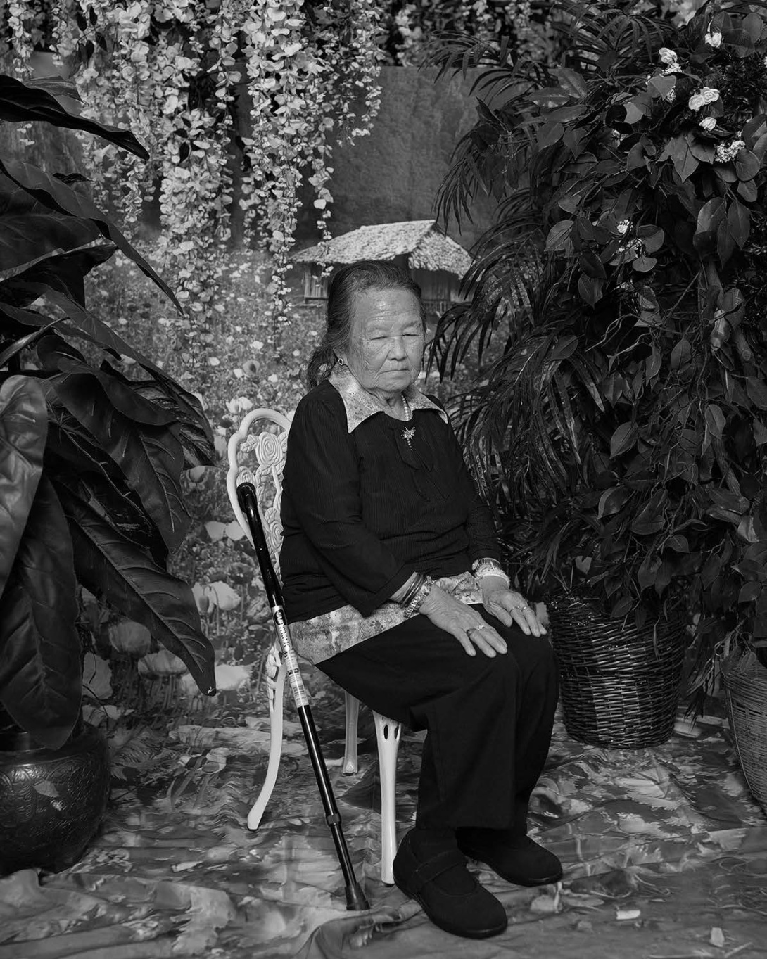 A black and white image of an older Asian woman. She sits on a chair in front of a printed country backdrop. She is surrounded by artificial flowers.