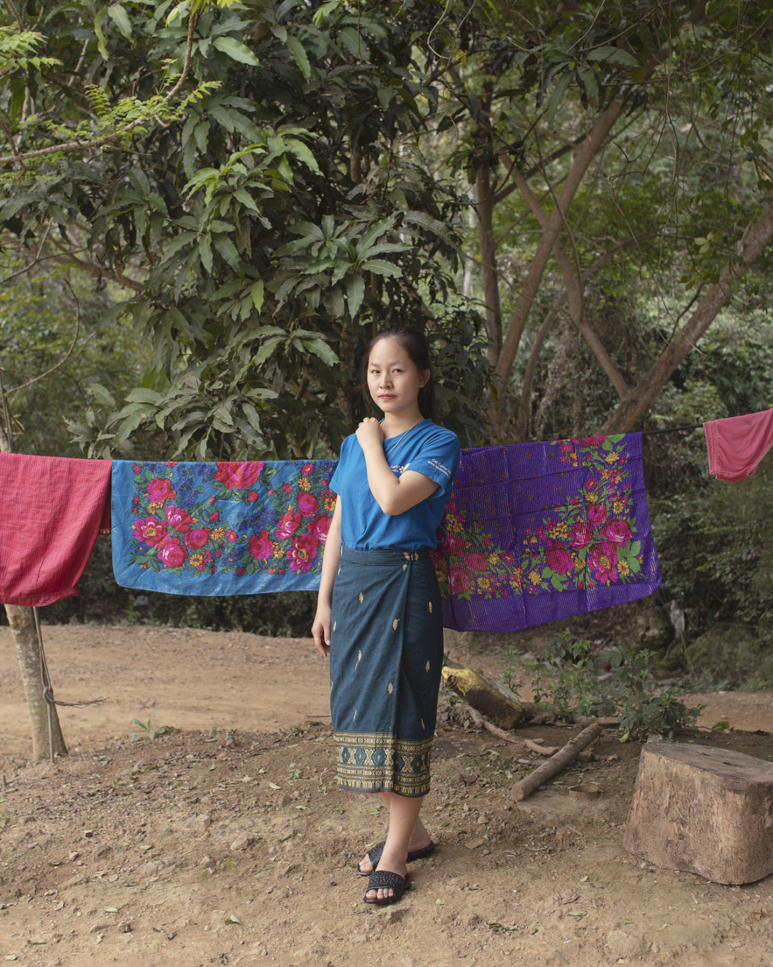 A young woman wears a blue shirt and a blue sarong with sandals. She stands in front of colorful floral laundry hanging on a line. Behind her are very full trees and a large tree stump.