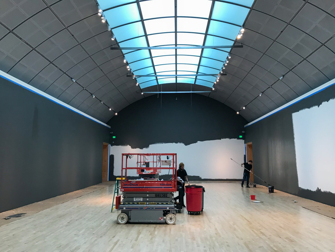 In a gallery with the walls two different colors are two people working—one is painting and the other is carrying something to a trash bin. 