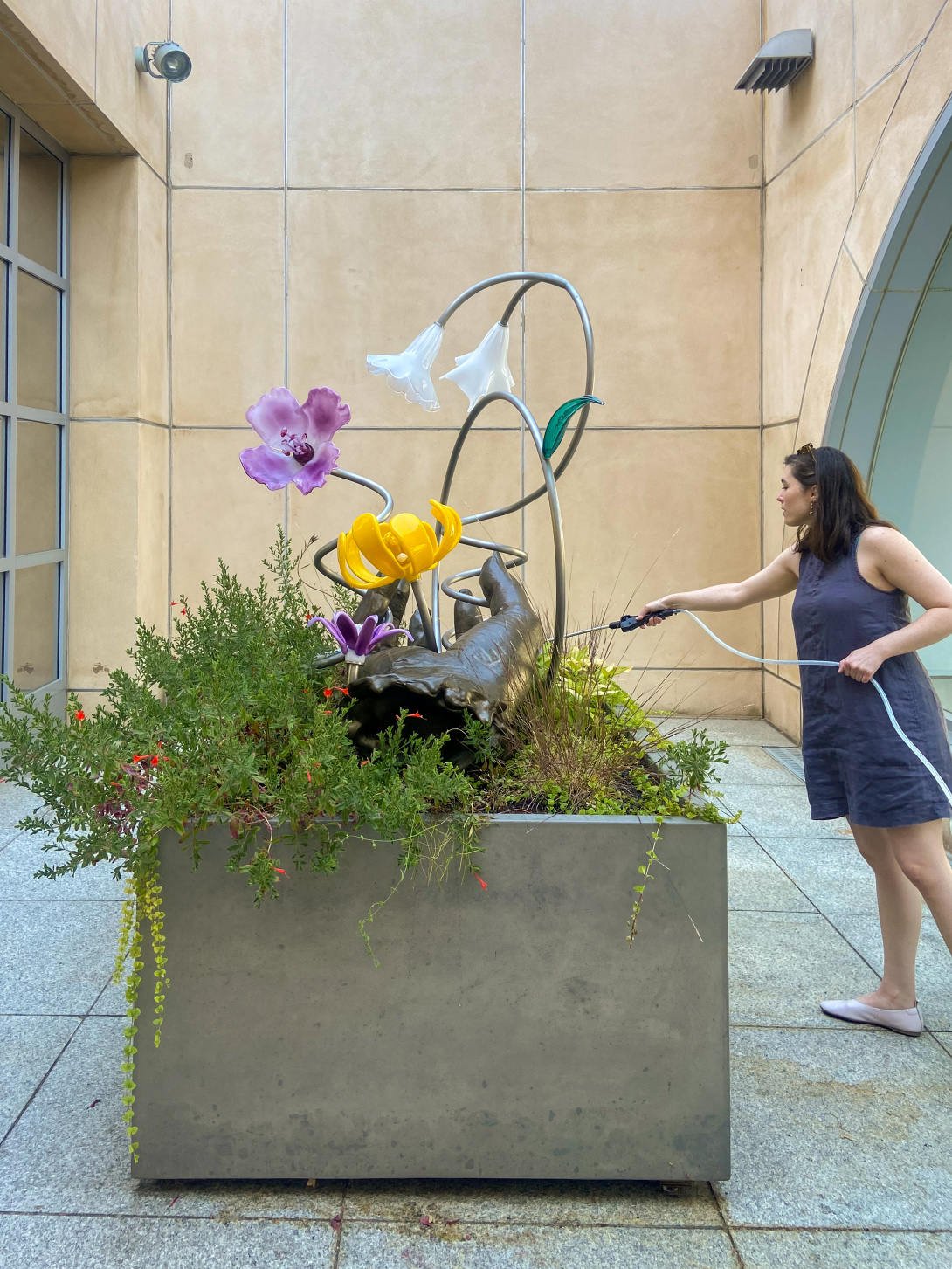 A woman is watering plants and flowers that surround a large iron and glass floral sculpture made of iron and glass.