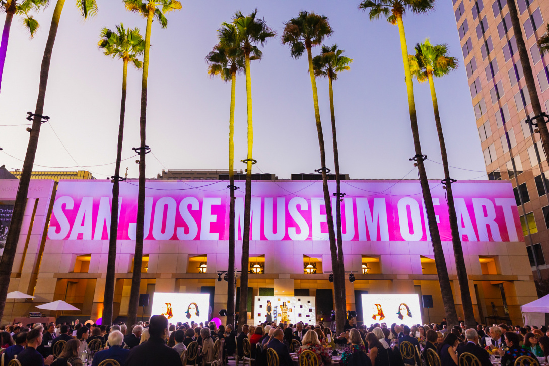 Evening gala at the San José Museum of Art with guests seated outdoors under tall palm trees, facing a stage and large screens in front of the museum's illuminated facade.