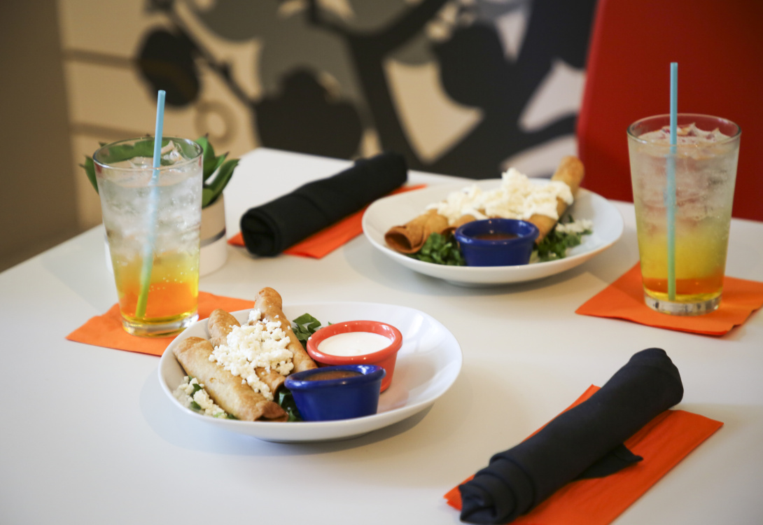 A table set with two plates of taquitos topped with cheese, accompanied by dipping sauces, drinks with blue straws, and black napkins on orange napkins.