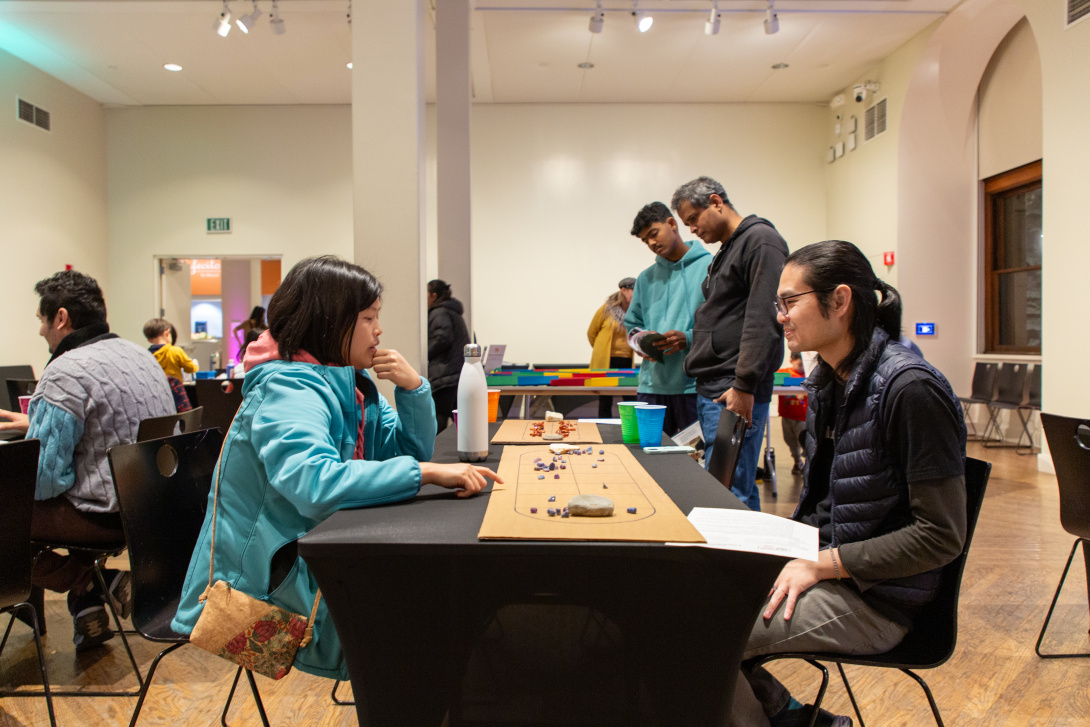 Two people sit across from each other with a game board between them. In the background are more people looking at games on other tables.