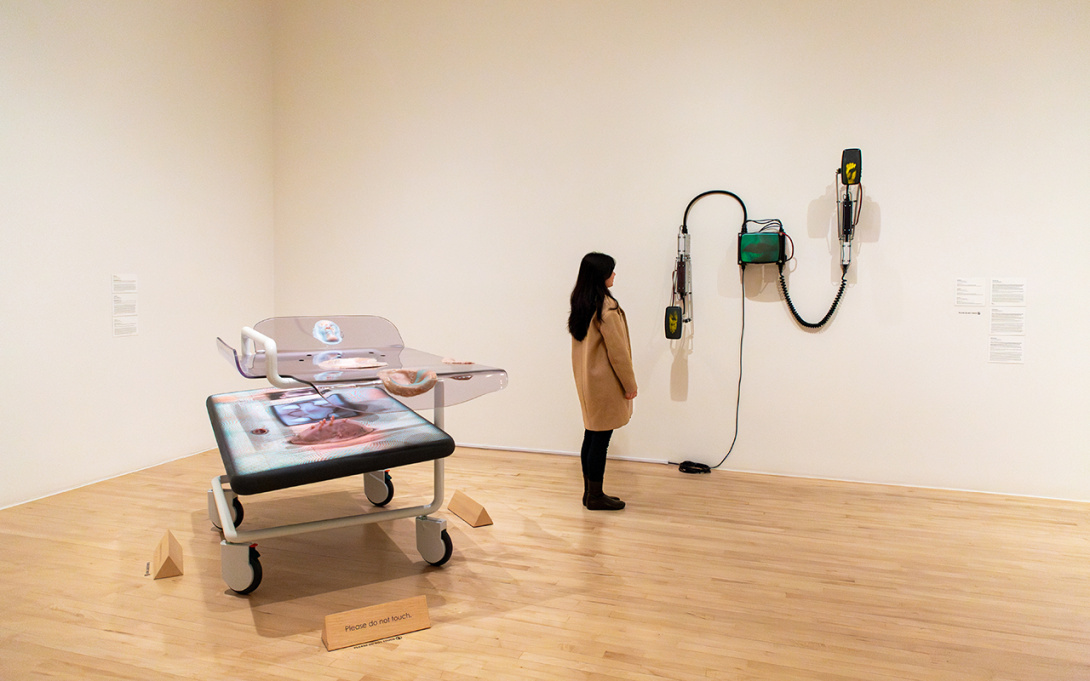 woman gazing at an electronic assemblage with a surreal hospital bed behind her