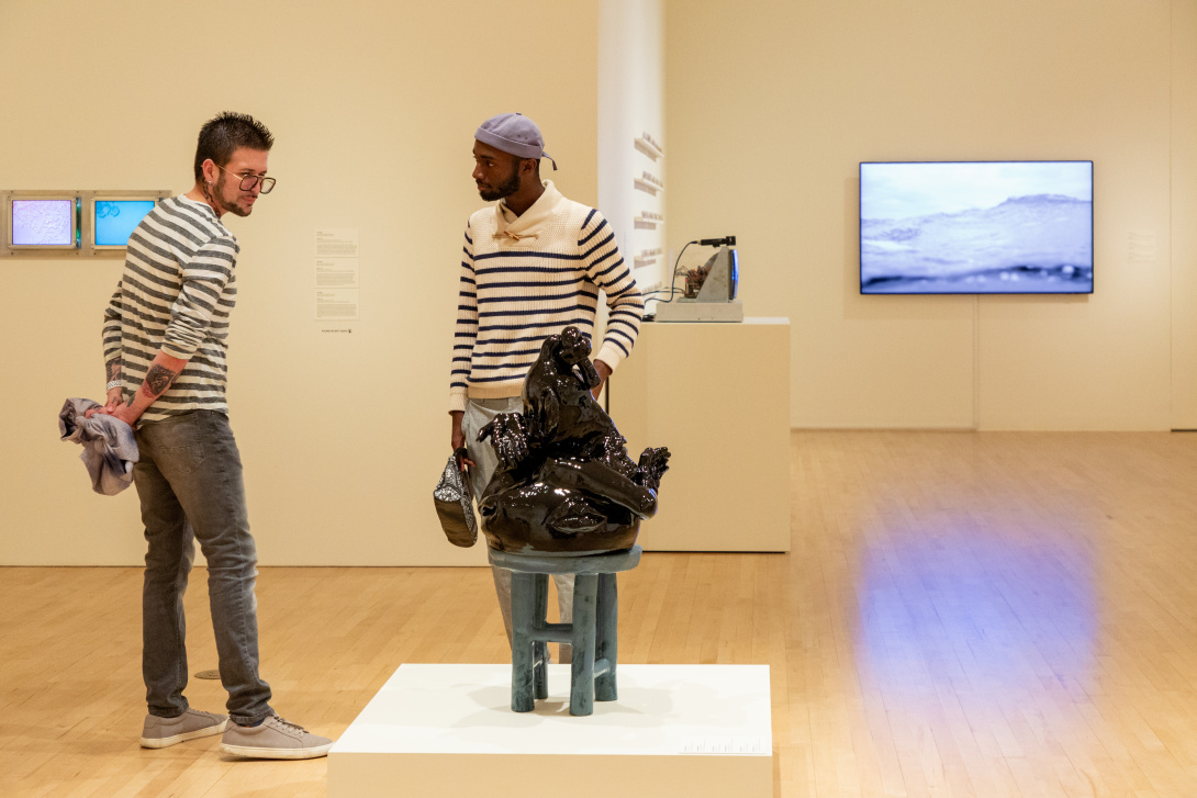 Two men wearing striped tops looking at an abstract ceramic sculpture on a stool