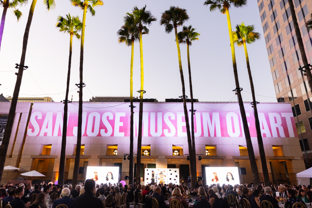 crowds of people dining underneath palm trees in front of a building with the San Jose Museum of Art banner