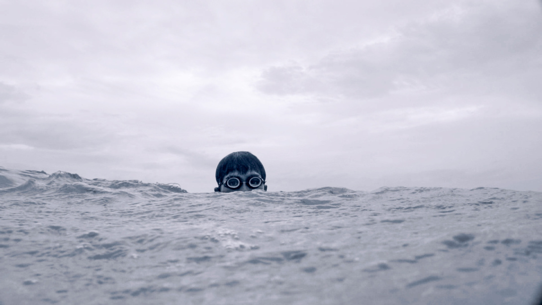 Person wearing swim goggles peers above a gray ocean surface beneath an overcast sky, with only their head visible above the water