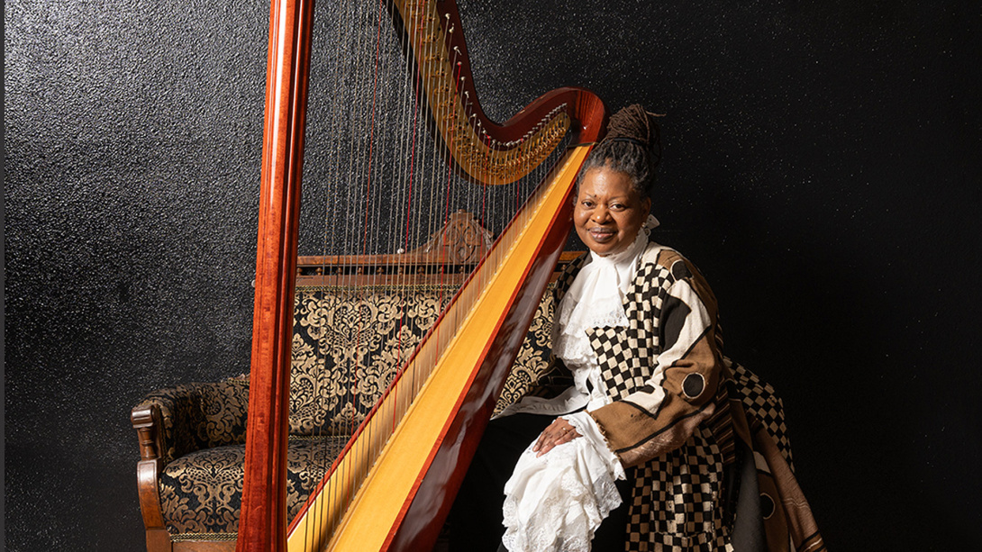 black woman with long dreads posed next to a harp in an up-do wearing a victorian-inspired dress with contemporary fabric 