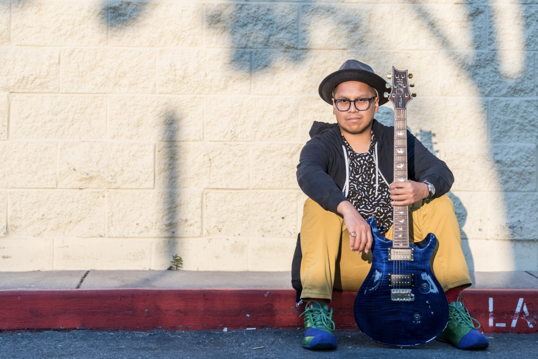 asian american man wearing a fedora holding a blue electric guitar seated on the curb