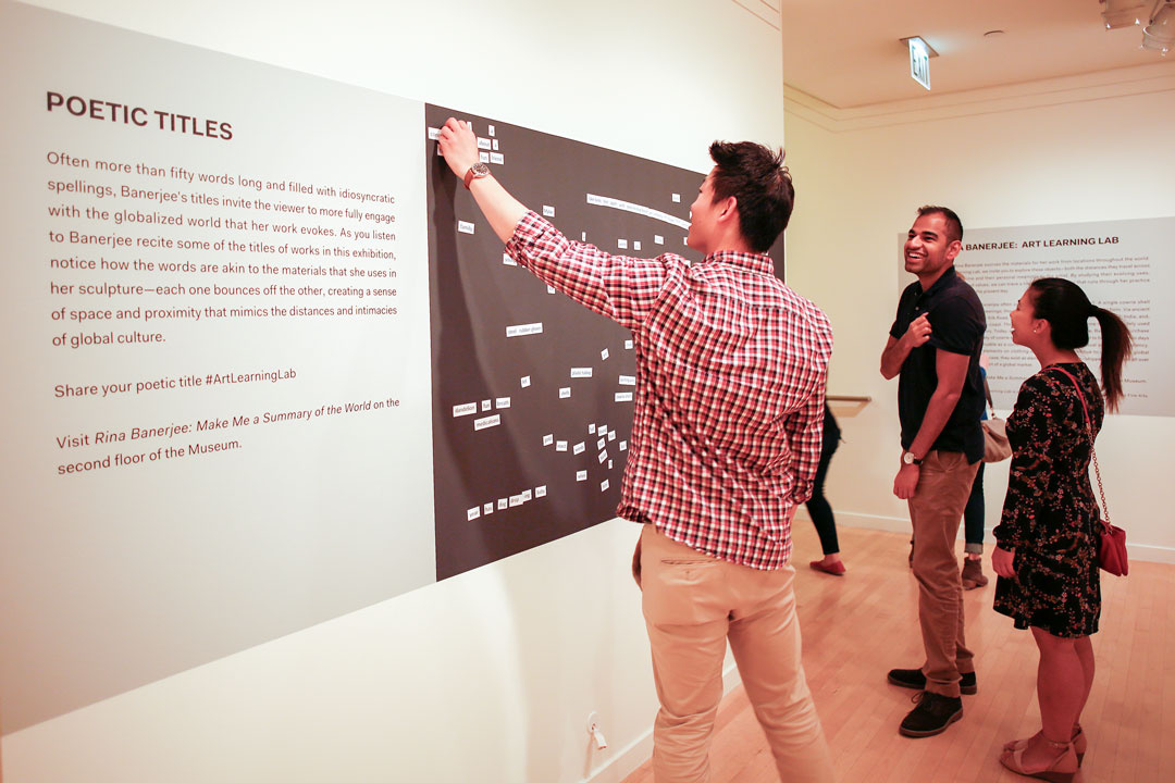 Three diverse visitors (two men and a woman) are enthusiastically engaged in a hands-on activity arranging magnetic poetry on a wall. The adjacent explanatory text identifies the project as “Poetic Titles” related to an exhibition by an artist Rina Banerjee.