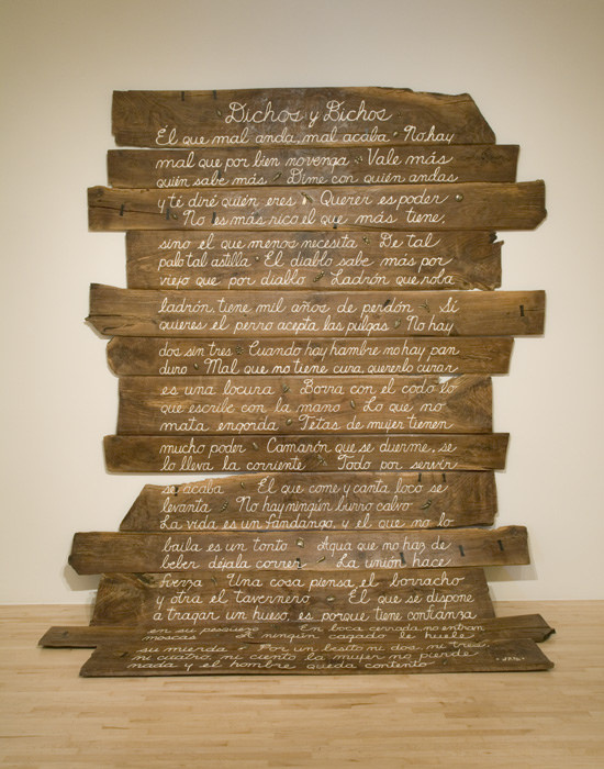 A long wooden sign that flows to the ground. The text is in Spanish and the pieces of wood put together are in different lengths. Also, the wooden pieces have some chips and dents on them—they resemble reclaimed wood. 