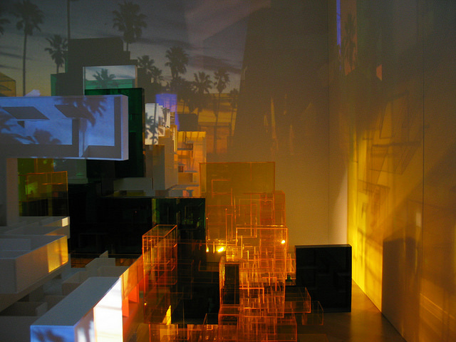 Light shining on orange colored Plexiglas boxes of varying heights in the center of a gallery casts elongated shadows on a nearby wall resembling sun beams reflected on skyscrapers. Behind the structure, a projected scene shows palm trees silhouetted against a sunset. 