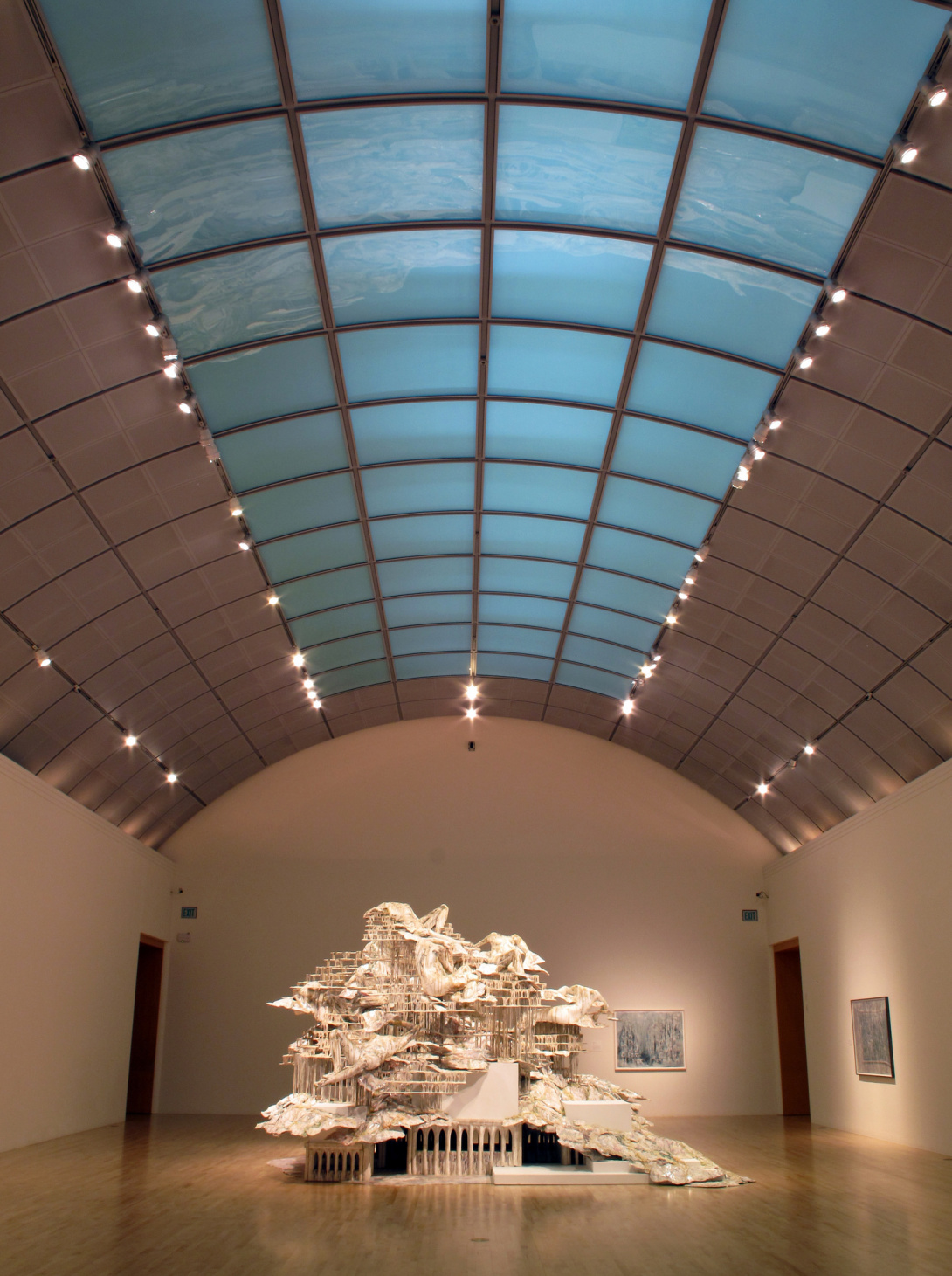 A large sculpture fills a gallery—like a cityscape with columns and boxes supporting the structure's height. It is covered in an unidentifiable white material is delicate and strong. It drips over the columns and boxes, appearing as roofs, treetops, clouds, and lounging oversized people.