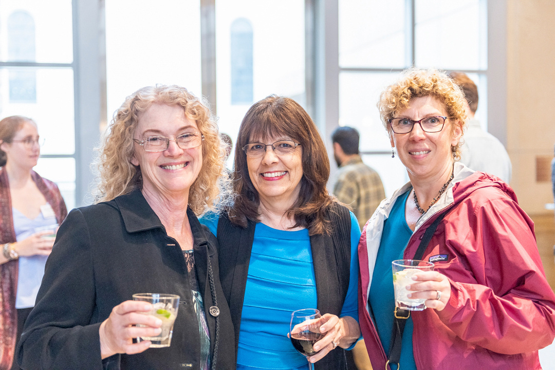 Three women pose for a photograph in an open museum space. They are holding beverages while people mill about in the background.
