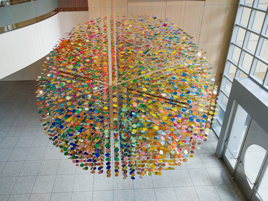 Thousands of colorful disks are suspended from the ceiling in a museum lobby. Altogether they make a large colorful sphere.