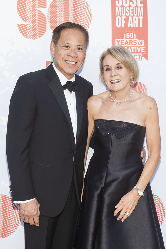 Older Asian man with older white blonde woman dressed in black suit and black evening gown respectively.