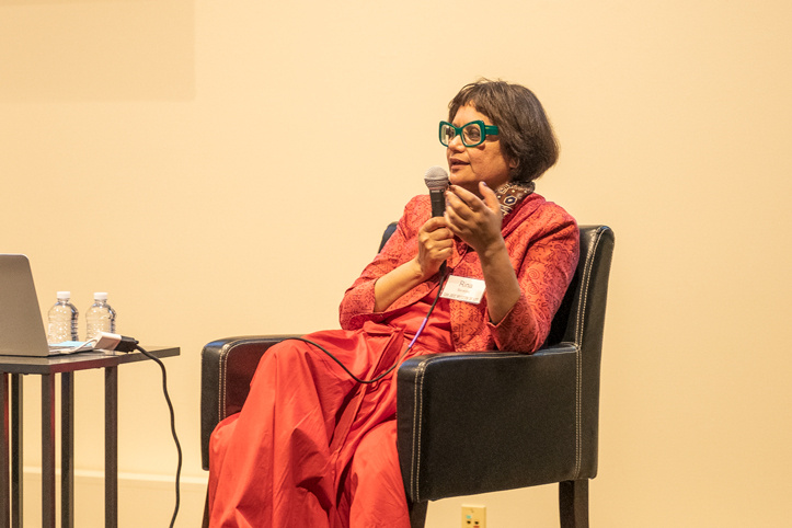 A guest speaker with green glasses in a red outfit holds a microphone and is seated next to a small table with a laptop and two small bottles of water. She is in the act of speaking.