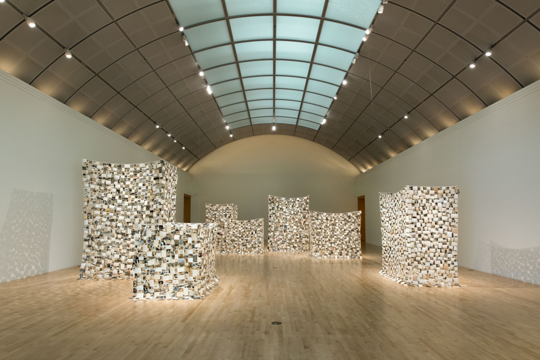 Spotlights showcase 7 large box-like structures in a long white gallery space with a high arched ceiling. Faintly visible strings from the ceiling hold each structure in formation, revealing their fragility and are precariously composed.