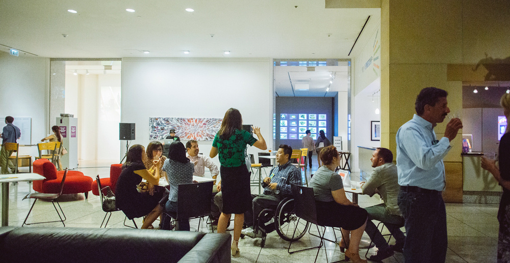 People socialize in a museum lounge, sitting at tables and engaging in conversation, with artwork and a well-lit gallery visible in the background.