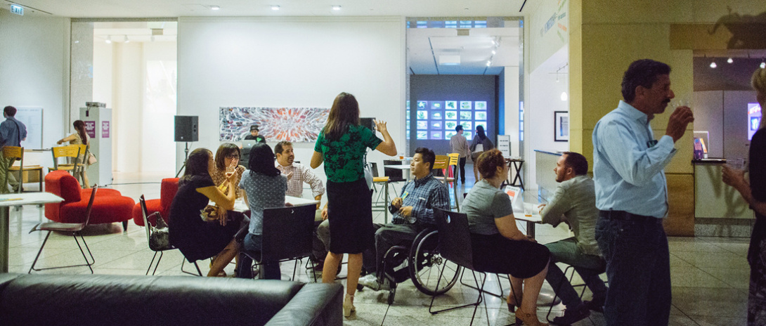 A museum lobby with a woman talking to a man in a wheelchair, others at a table, and a man holding a drink. A large painting and open galleries are in the background.