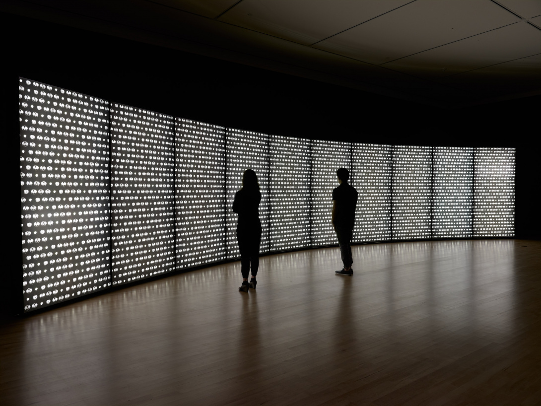 A large panel wall of lights curves in a dark installation space. The 10 panels have a translucent quality to them—the lights are in rows resembling dots and circles. Two people stand in front of the panels, observing the glowing installation.