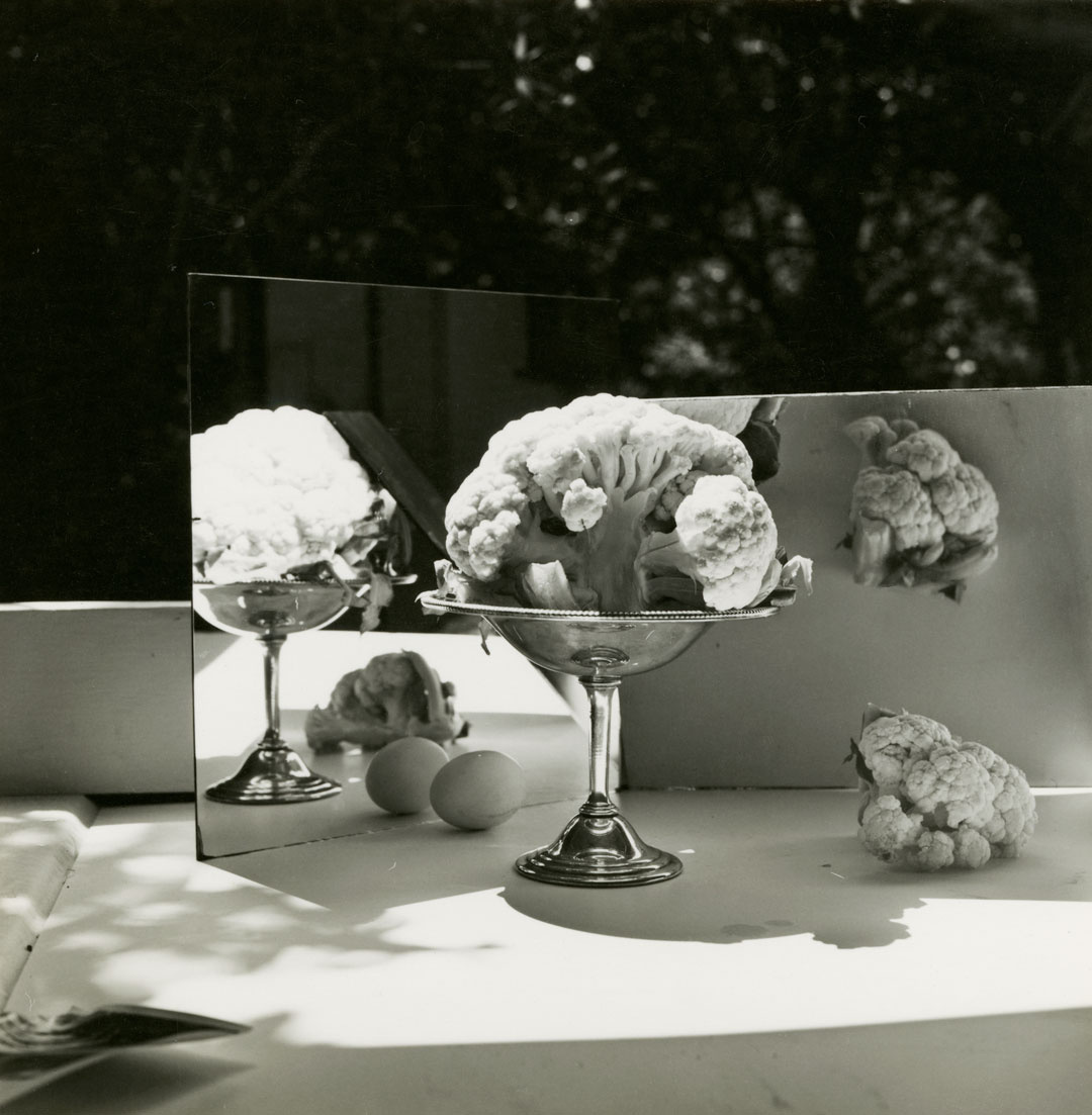 A black and white photograph of a cauliflower head in a silver goblet on a shelf or outdoor bench. Two mirrors sit behind it at skewed angles, revealing the contrasting shape of its far side and introduces new otherwise invisible elements including two eggs at its base. 