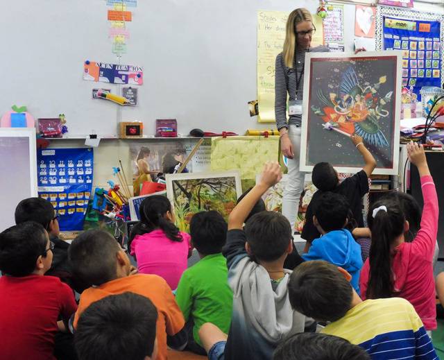 An adult instructor stands in front of colorful paper signs and decorations. She faces a classroom full of young children, presumably students. The children sit on the ground. Some raise their hands eagerly. The instructor holds a large poster board. 