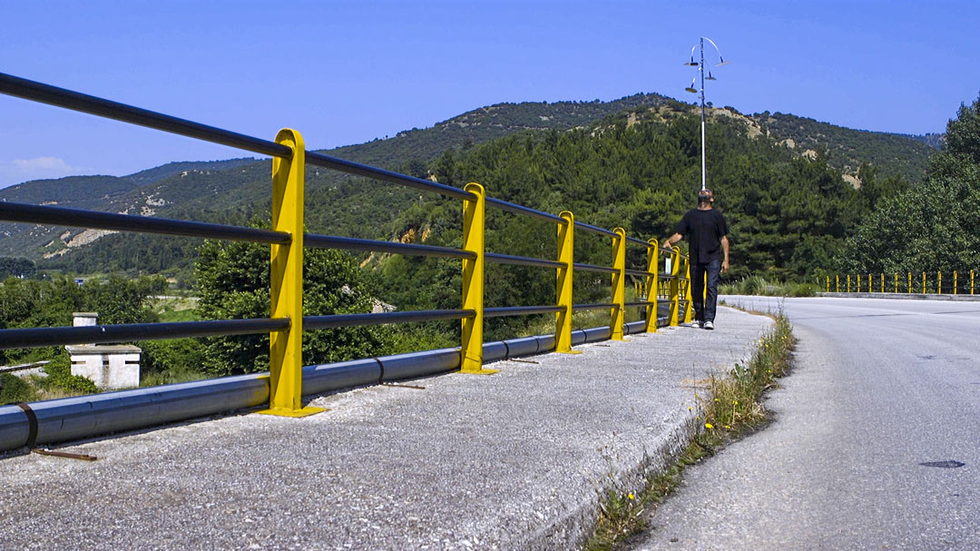 In the distance, a black man walks forward, holding onto a yellow steel sidewalk railing as he at a clear blue sky. Behind him is a mountain filled with green trees. Next to the sidewalk he is on is a street. Suburbs are below, in the foreground.