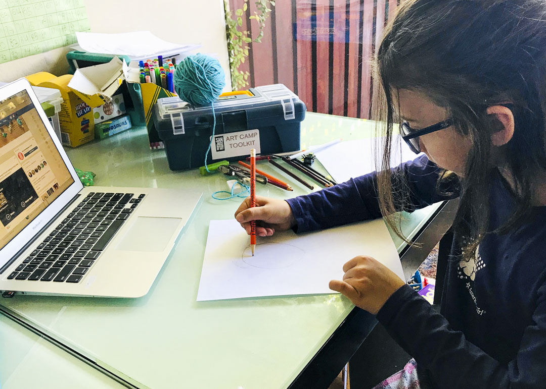A young girl with long hair wearing glasses sits at a desk drawing with an orange colored pencil on paper. Within her view is an open laptop, in the background resides a variety of colored pencils, an art camp toolkit, and unspooled turquoise yarn.