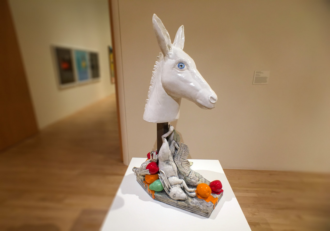 Glazed still-life sculpture of a blue-eyed, white donkey head on a stake, looking up. A grey rabbit, birds, and dripping fruit—including apples, oranges, and a pear—are arranged haphazardly at the base, suggesting they are dead.