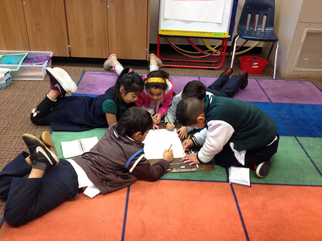 Five young children lay on a carpeted surface in a classroom drawing with pencils on a large piece of paper. The children work together and appear concentrated on their drawing as they lay in a circular formation around the edges of the paper. 