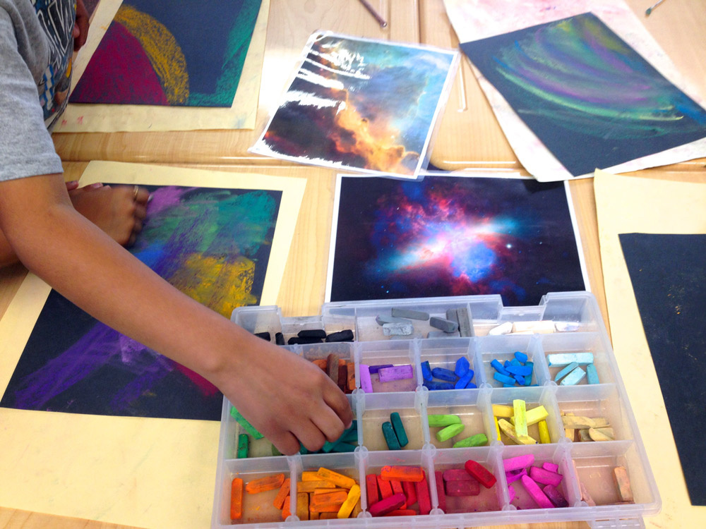 An aerial perspective of a wooden table with art supplies. A child's hand reaches across the table to obtain an oil pastel stick from a plastic container organized by color. On the table are drawings on black construction paper of outer space imagery. 