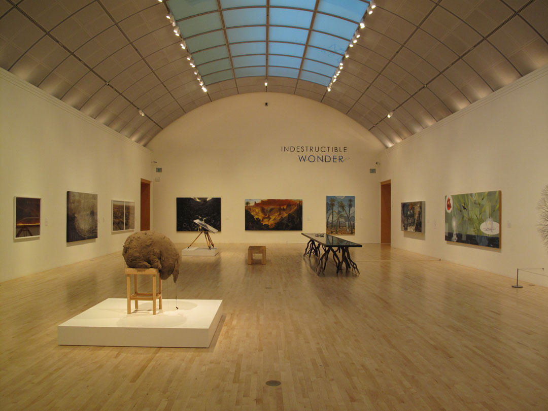A photograph of an art gallery with multiple paintings on the walls with four additional sculptures in the middle of the room. An arched celling with glass panes runs through the ceiling.