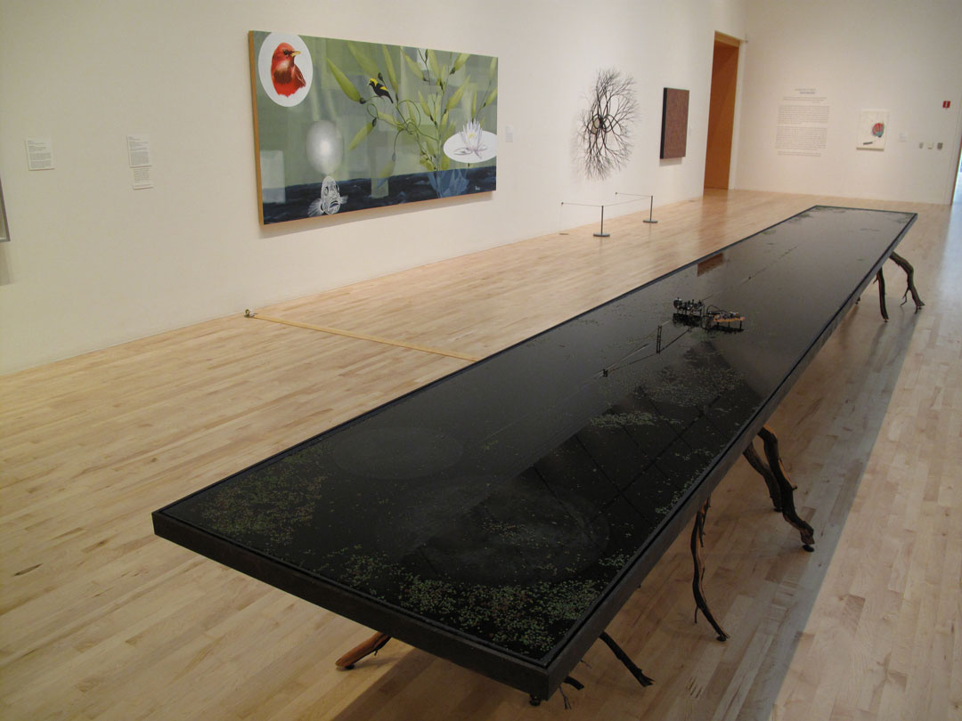 A long rectangular black table that appears to have water in it with greenery inside it. It has multiple legs resembling tree branches. On the wall is a painting of birds, along with a sculpture that sticks out which also resembles tree branches.