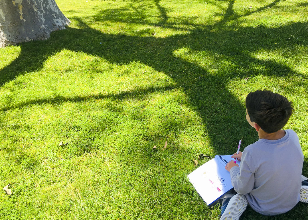 A young child sits on a grassy field on a sunny day facing a tree in the distance. The child holds a spiraled notebook in their lap and a marker in their right hand. 