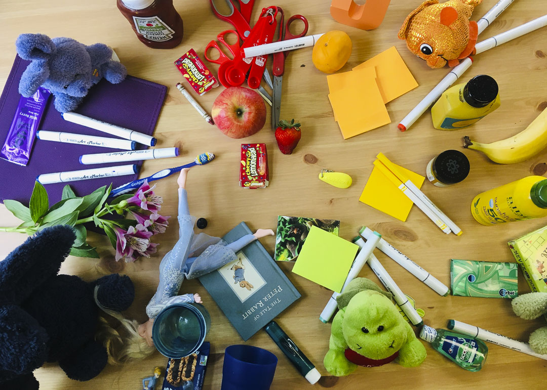 A wooden table viewed from an aerial perspective with miscellaneous items arranged in groups based on color. Items include; an apple, a strawberry, a banana, post-its, a packet of bubblegum, markers, a crayon, stuffed animals, scissors, a book, etc. 