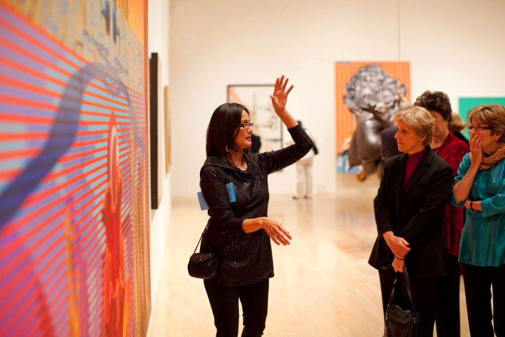 A docent or interpreter stands in a museum gallery in front of a very colorful painting. She holds her hands aloft, addressing a group of women. Colorful paintings hang on the walls and a large bronze sculpture hangs in the background, out of focus.  