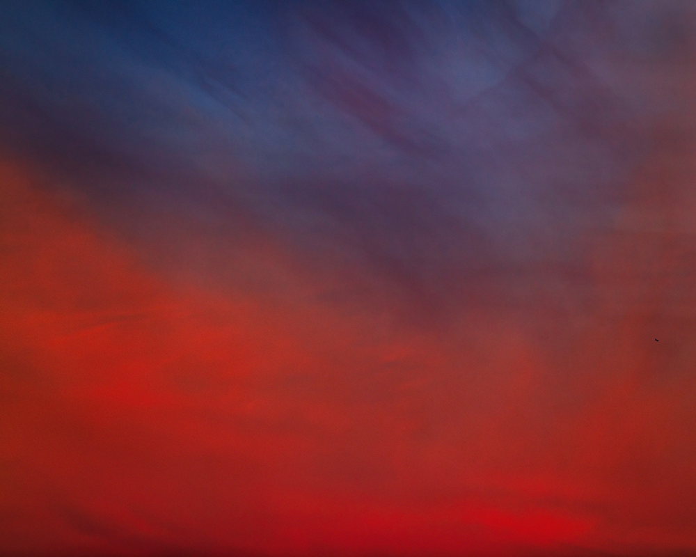 A sky of bright red rising to deep blue, with light and shadows casting rich contrast. Barely visible on the right hand edge where the red and blue meet to form purple is a small black speck: a drone.
