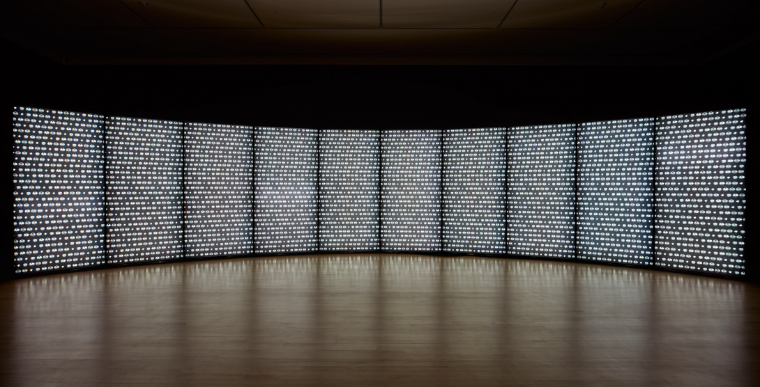 A large panel wall of glowing lights curves in a dark installation space. The 10 panels have a translucent quality to them—the lights are in rows resembling dots and circles. The white light is reflected on the glossy wooden floor.